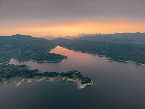 Aerial view of Wadaslintang Reservoir at sunset with calm water reflecting the orange sky, surrounded by lush green hills and dense vegetation in Wadaslintang, Central Java, Indonesia.