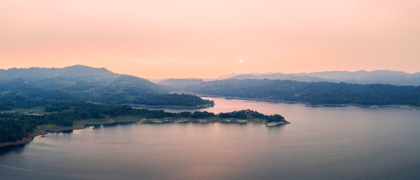 Aerial view of Wadaslintang Reservoir at sunset with calm water reflecting the soft light and rolling hills in the background in Wadaslintang, Central Java, Indonesia.