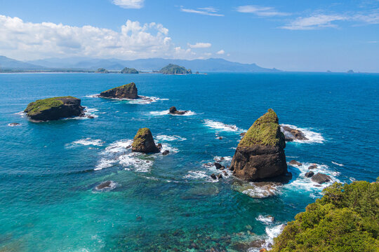 Aerial view of Papuma Beach with dramatic rock formations rising from turquoise waters and lush green vegetation under a clear blue sky in Kecamatan Wuluhan, Jawa Timur, Indonesia.