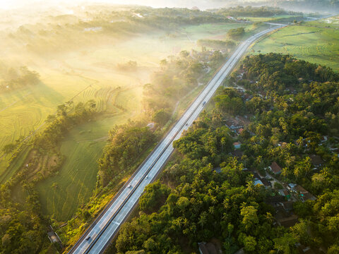 Aerial view of the Kemetul highway winding through lush rice fields and dense tropical greenery under the soft morning mist in Susukan, Central Java, Indonesia.