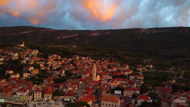 Aerial view of Jelsa with its historic stone rooftops, church bell tower, and harbor illuminated by the warm glow of sunset in Jelsa, Split-Dalmatia County, Croatia.