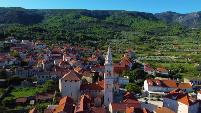 Aerial view of the historic town of Jelsa with its prominent church tower, terracotta rooftops, and surrounding lush green hills in Jelsa, Split-Dalmatia County, Croatia.