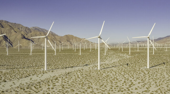 Aerial view of the San Gorgonio Pass Wind Farm with numerous white wind turbines standing in the arid desert landscape under a clear blue sky in Palm Desert, California, United States.