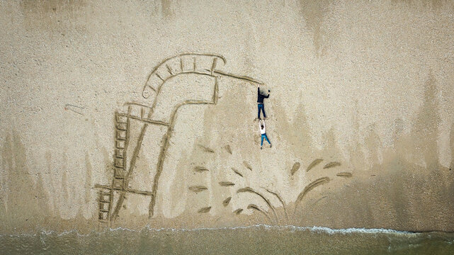 Aerial view of two people interacting with a creative sand drawing of a diving board and water splash on the sandy beach, Damgan, Brittany, France.