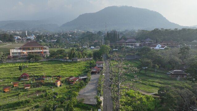 Aerial view of Trawas featuring lush green rice terraces, small wooden structures, a winding road, and distant mountains in Trawas, East Java, Indonesia.