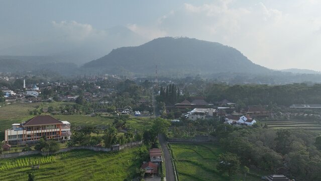 Aerial view of Trawas with lush green rice terraces, small buildings, and a large mountain in the background under a hazy sky in Trawas, East Java, Indonesia.