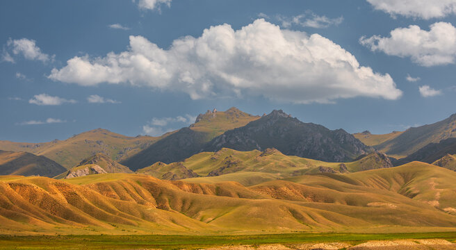 View of the rolling grassy hills and rugged mountain peaks of Sarybulak under a bright blue sky with scattered white clouds in Sarybulak, Osh Region, Kyrgyzstan.