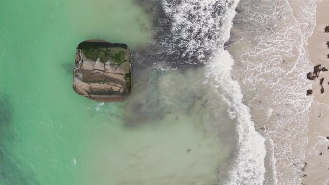 Aerial view of Haukland Beach with turquoise water and waves crashing against a large mossy rock on the sandy shore in Leknes, Nordland, Norway.