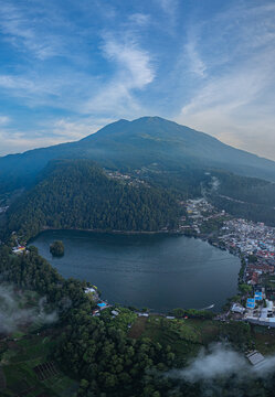 Aerial view of Sarangan Lake surrounded by lush green forests and the majestic Mount Lawu with a small town nestled along the shore in Plaosan, East Java, Indonesia.