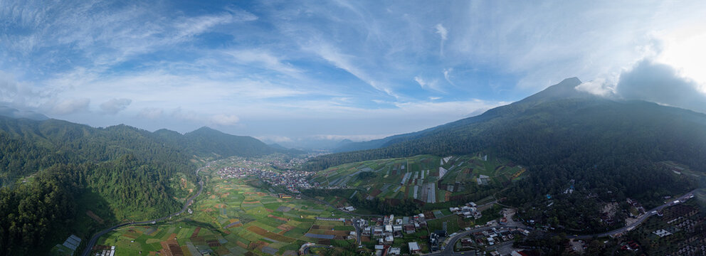 Aerial view of Sarangan lake area with lush agricultural fields, small town, and surrounding mountains under a blue sky in Plaosan, East Java, Indonesia.