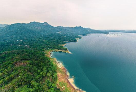 Aerial view of Wadaslintang Reservoir surrounded by lush green hills and dense forest with calm blue water and agricultural plots in Wadaslintang, Central Java, Indonesia.
