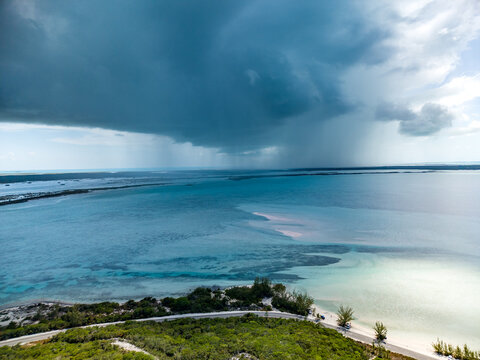Aerial view of the coastal landscape of Rolleville with turquoise waters, lush green vegetation, and a dramatic storm cloud bringing rain over the sea in Rolleville, Exuma, The Bahamas.