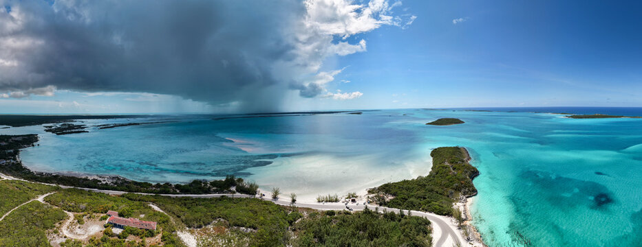 Aerial view of the coastal landscape of Rolleville, featuring turquoise waters, a sandy beach, lush vegetation, and a storm cloud gathering over the horizon in Rolleville, Exuma, The Bahamas.