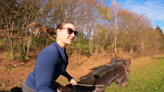 SELFIE: Young woman rides a dark brown horse across green meadow, following chestnut mare galloping ahead. Horseback riding through countryside in the embrace of colorful autumn forest under blue sky