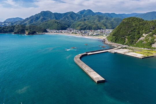Aerial view of the coastal town of Matsuzaki, featuring a concrete breakwater extending into the turquoise sea, backed by lush green mountains in Matsuzaki, Shizuoka, Japan.