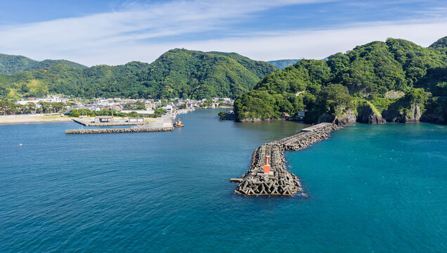 Aerial view of the coastal harbor in Matsuzaki featuring a breakwater with a red lighthouse, calm blue water, and lush green hills in the background in Matsuzaki, Shizuoka, Japan.