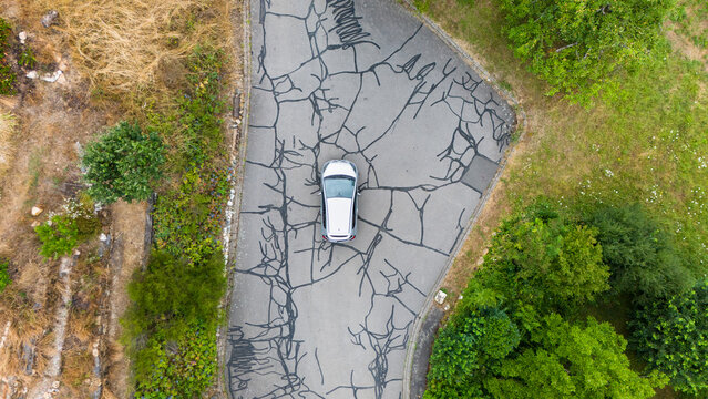 Aerial view of a car parked on an asphalt road with extensive tar-filled cracks, surrounded by green trees and dry grass in Trier-Saarburg, Rhineland-Palatinate, Germany.