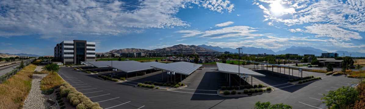 Aerial view of a modern office building with extensive solar panel parking structures, surrounded by mountain vistas under a bright blue sky in Lehi, Utah, United States.