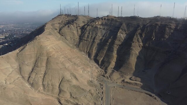 Aerial view of the Morro Solar hill in Chorrillos, featuring communication towers on the summit, rugged desert terrain, and winding paths overlooking the urban landscape of Lima, Lima, Peru.