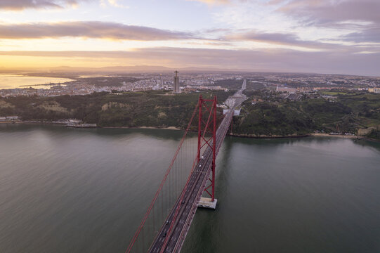Aerial view of Ponte 25 de Abril suspension bridge spanning the Tagus River at sunset with the Sanctuary of Christ the King statue visible in the distance Lisbon, Lisbon, Portugal.