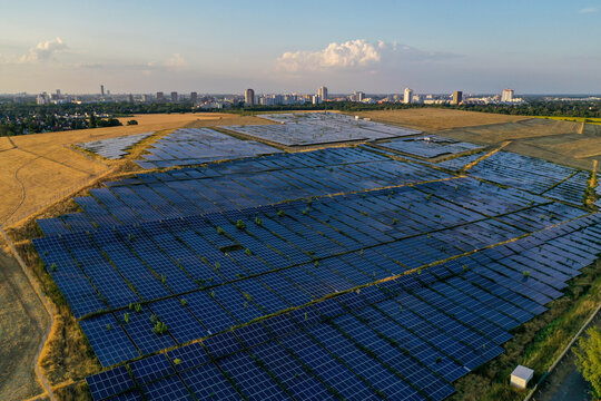 Aerial view of a solar farm with rows of photovoltaic panels, with the Berlin skyline and TV tower visible in the distance under a clear sky at golden hour, Schonefeld, Brandenburg, Germany.