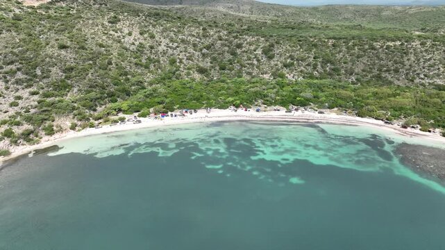 Aerial view of Playa Blanca with turquoise water, lush green hills, and cars parked along the sandy shore in Azua, Azua Province, Dominican Republic.