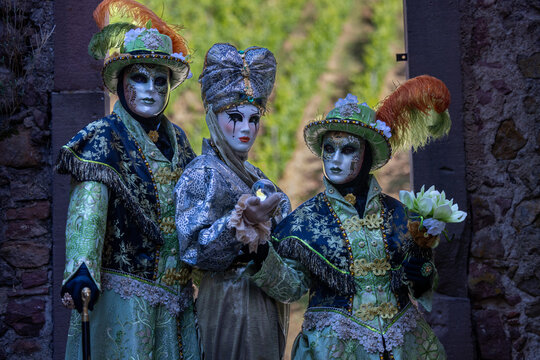Riquewihr, France - 04 July 2026: View of three people wearing elaborate Venetian masks and ornate costumes standing in a stone archway.