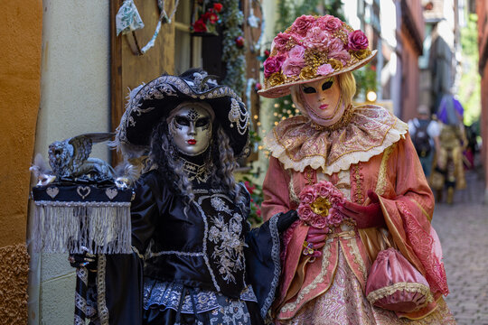 Riquewihr, France - 04 July 2026: View of two people in elaborate Venetian carnival costumes and masks standing in a narrow historic street.