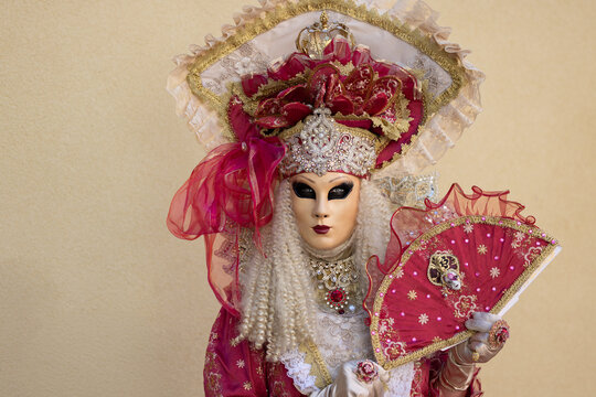 Riquewihr, France - 04 July 2026: View of a person wearing an elaborate Venetian carnival mask and a red and gold ornate costume while holding a matching decorative fan.
