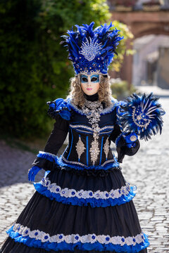 Riquewihr, France - 04 July 2026: View of a person wearing an elaborate blue and black Venetian-style mask and costume with intricate lace and feathers, standing on a cobblestone street.