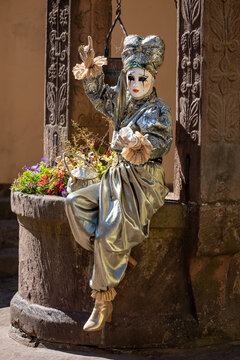 Riquewihr, France - 04 July 2026: View of a person in an elaborate silver and gold carnival costume sitting on the edge of an old stone well decorated with colorful flowers.