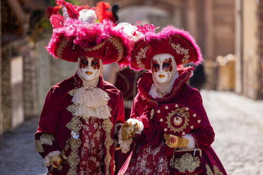 Riquewihr, France - 04 July 2026: View of two people in elaborate red and gold Venetian carnival costumes and masks walking on a historic cobblestone street in Riquewihr.