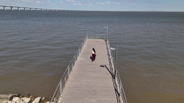 Aerial view of a woman walking along the wooden pier at Parque das Nacoes with the Vasco da Gama Bridge in the background in Lisbon, Lisbon, Portugal.