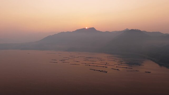 Aerial view of Wadaslintang Reservoir at sunrise with misty mountains and fish farming cages in the water in Wadaslintang, Central Java, Indonesia.