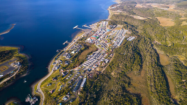 Aerial view of the town of Puerto Williams nestled along the Beagle Channel, surrounded by dense forests and rugged hills in Puerto Williams, Magallanes, Chile.