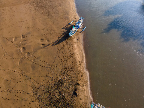 Aerial view of a traditional outrigger boat moored on the sandy riverbank with visible footprints in the sand near the water in Kebumen, Central Java, Indonesia.