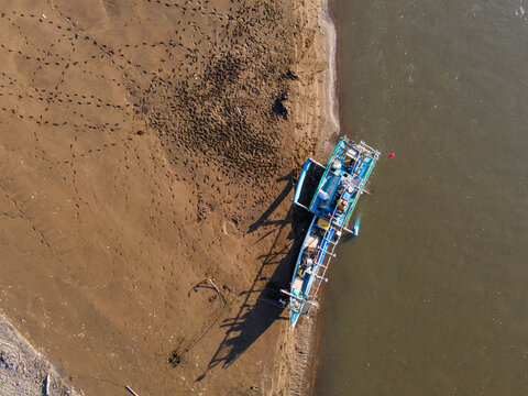Aerial view of a traditional fishing boat moored on the sandy beach with visible footprints and calm coastal waters in Kebumen, Central Java, Indonesia.