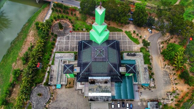 Aerial view of the Great Mosque of Panyabungan with its green tower and dark roof, surrounded by trees, a river, and paved courtyards in Panyabungan, North Sumatra, Indonesia.