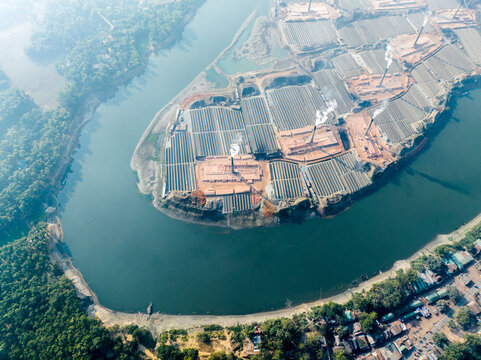 Aerial view of brick kilns with smoking chimneys and rows of bricks situated along a river bend in Tangail, Dhaka Division, Bangladesh.
