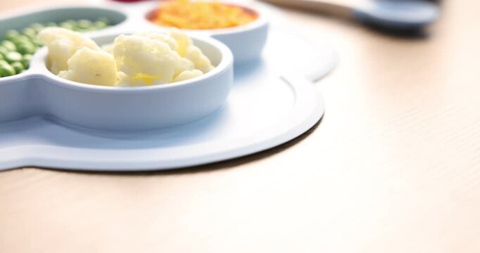 Baby tableware. Section plate with food and cutlery on wooden table, closeup. Camera moving right