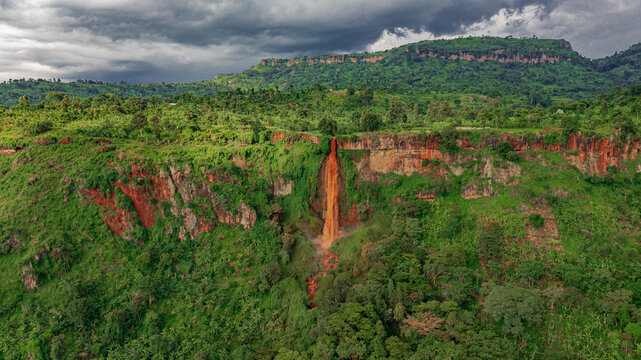 Aerial view of Sisiyi Falls cascading down a reddish-brown cliff face surrounded by lush green vegetation and rolling hills under a cloudy sky in Buligenyi, Mkoa wa Mashariki, Uganda.