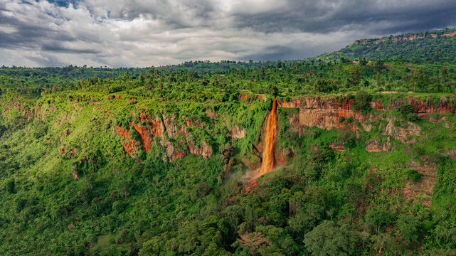 Aerial view of Sisiyi Falls cascading down a lush green cliffside with vibrant red rock formations under a cloudy sky in Buligenyi, Mkoa wa Mashariki, Uganda.