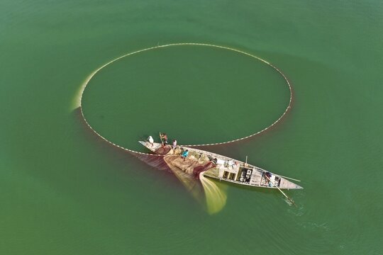 Aerial view of fishermen in a wooden boat casting a large circular fishing net into the calm green water in Gaibandha, Rangpur Division, Bangladesh.