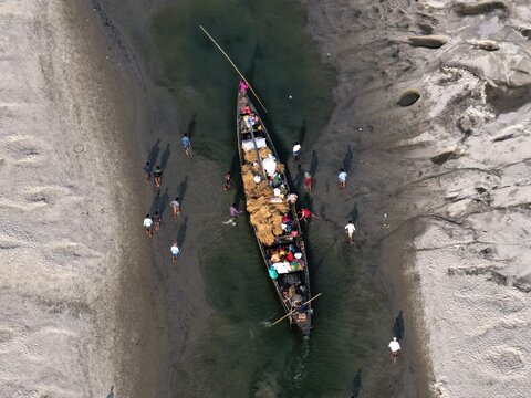 Aerial view of a traditional wooden boat loaded with straw in a shallow sandy riverbed with people walking along the banks in Gaibandha, Rangpur Division, Bangladesh.