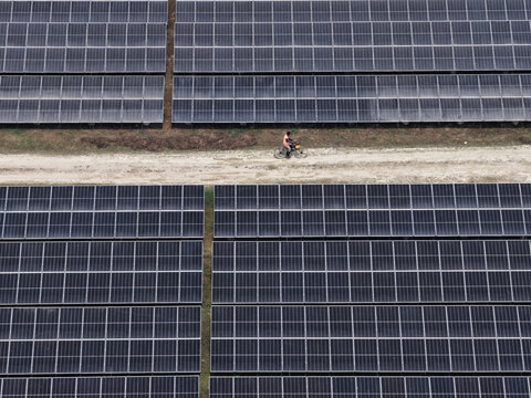 Aerial view of a man riding a bicycle on a dirt path between rows of solar panels at a solar power plant in Gaibandha, Rangpur Division, Bangladesh.