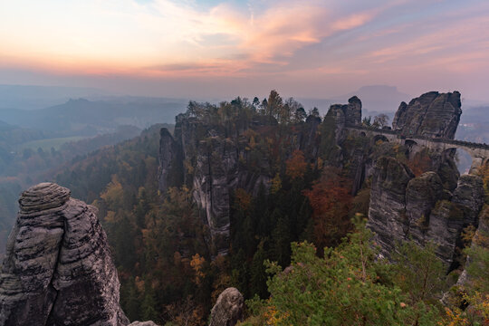 View of Bastei Bridge surrounded by dramatic sandstone rock formations and autumn forest under a soft sunset sky in Germany.
