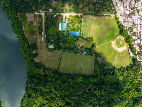 Aerial view of Casa Club Colegio San Judas Tadeo featuring a soccer field and baseball diamond surrounded by lush trees and a river in Bajos de Haina, San Cristobal Provinc, Dominican Republic.
