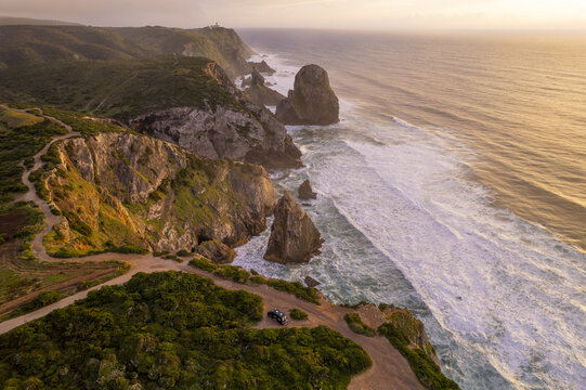 Aerial view of Cape Roca featuring rugged cliffs meeting the Atlantic Ocean at sunset with a winding path and a camper van parked on the cliff edge in Colares, Lisboa, Portugal.