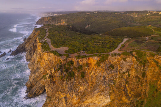 Aerial view of Cape Roca featuring rugged cliffs overlooking the Atlantic Ocean with waves crashing against the rocks during golden hour in Colares, Lisboa, Portugal.