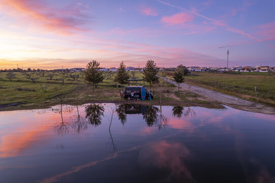 Aerial view of a camper van parked by a pond at sunset with vibrant pink and orange sky reflections in the water, surrounded by trees and rural fields in Oriola, Distrito de Evora, Portugal.